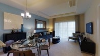 Located at 1 Park Lane Boulevard in Cherry Hill, New Jersey, this single-floor residence offers a thoughtfully designed open-concept layout that seamlessly blends living and dining spaces. The living area is anchored by plush carpeting and a tray ceiling that adds architectural depth and dimension to the room. A large sliding glass door floods the space with natural light while providing easy access to the outdoors, complemented by patterned drape panels that add a decorative touch.

The dining area features a sleek glass-top table with contemporary seating, positioned beneath a chandelier that serves as an elegant focal point. A large framed mirror mounted on a richly toned accent wall enhances the sense of openness and reflects light throughout the room. The living space is furnished with a deep navy sofa accented with teal and gold throw pillows, a round ottoman coffee table, and a cushioned accent chair, creating a cohesive and inviting atmosphere.

Neutral wall tones are offset by a striking blue-gray accent wall, giving the space a modern yet warm character. The overall ambiance is sophisticated and welcoming, with carefully chosen décor elements — including floral arrangements and layered lighting — that contribute to a refined residential aesthetic throughout.