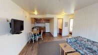 This single-floor suite is designed for functional, self-sufficient living, combining a living area, kitchen, dining space, and bedroom within a practical open layout. The main living area features wood-look vinyl flooring and is furnished with a patterned sofa, a low coffee table, and a wall-mounted flat-screen television, creating a comfortable and straightforward lounge space. A small area rug anchors the seating zone, visually separating it from the rest of the open floor plan.

Adjacent to the living area, a compact dining nook accommodates a round wooden table with white chairs, making the most of the available space. The kitchen flows directly from the dining area and is equipped with warm oak cabinetry — both upper and lower — dark granite-style laminate countertops, a stainless steel single-basin sink, a white refrigerator, and a gas range, providing all the essentials for meal preparation. The kitchen's L-shaped counter configuration maximizes workspace efficiently.

A hallway visible from the main area leads to a separate bedroom, offering a degree of privacy. The textured ceiling and neutral white walls throughout give the suite a clean, no-frills appearance well-suited to practical, everyday use.
