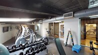 This fitness center offers a comprehensive workout environment designed to accommodate a wide range of exercise needs. The space features an industrial aesthetic, with an exposed dark ceiling revealing structural beams, ductwork, and ventilation systems that lend the facility a functional, no-frills character. Rubber flooring throughout provides both safety and durability across all workout zones.

The main cardio area is lined with a row of treadmills, elliptical machines, and stationary bikes, offering ample options for aerobic training. Adjacent to this, a dedicated strength training section houses a multi-station cable machine, adjustable weight benches, and a full rack of dumbbells in varying weights, neatly organized for easy access. A colorful kettlebell rack and step platform add versatility for functional fitness routines.

A separate alcove provides a more intimate workout zone, complete with a freestanding heavy punching bag, a speed bag, stability balls, and additional dumbbells — ideal for boxing drills, core work, and supplementary training. A wall-mounted television is positioned in this area for added convenience.

Large mirrors are strategically placed to maximize the sense of space and allow users to monitor their form. A glass-paneled room adjoining the main floor appears to serve as a designated stretch or studio area. The facility also includes clearly marked, separate restrooms conveniently located within the space.