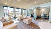 Located at 13325 Station Rail Way in Middletown, Kentucky, this single-floor amenity space combines modern design with functional comfort. The main lounge area is bathed in natural light streaming through floor-to-ceiling windows, offering expansive views of the surrounding Middletown skyline and streetscape. Plush cream-toned seating arrangements, patterned carpet tiles, and elegant marble-topped tables create an inviting atmosphere for relaxation or casual gathering.

A distinctive slatted wood ceiling runs throughout the lounge, lending warmth and architectural character to the space. A glass-enclosed private workroom with dark desks and ergonomic chairs provides a quiet, professional setting, while a beverage station with stylish cabinetry adds a refined, hospitality-inspired touch nearby. Bookcase displays set against decorative floral wallcovering add a library-like charm to one corner of the lounge.

A glass-walled fitness center is accessible from the main corridor, equipped with cardio and strength training equipment and filled with natural light. Dramatic globe pendant chandeliers hang over a mezzanine-level overlook, creating a striking focal point. Throughout, the space balances sophisticated finishes with an open, welcoming design suited for both productivity and leisure.