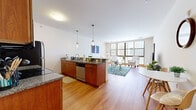 This thoughtfully designed one-bedroom, one-bathroom unit offers a comfortable and functional single-floor layout with approximately 790 square feet of living space. The open-concept kitchen and living area forms the heart of the unit, anchored by warm cherry wood cabinetry, granite countertops, and stainless steel appliances. A peninsula with bar seating and pendant lighting above creates a natural divide between the kitchen and living area, while hardwood flooring flows throughout, enhancing the sense of continuity and warmth. The living room is bathed in natural light from large floor-to-ceiling windows, which frame views of the surrounding urban streetscape and provide a bright, airy ambiance. A dedicated dining nook adds additional functionality to the main living space. The primary bedroom is a comfortable retreat, featuring soft carpeting, expansive windows overlooking an outdoor seating area, and ample space for full-size furnishings. The full bathroom is finished with tile flooring, a granite-topped vanity with warm wood cabinetry, and a full tub-shower combination. A dedicated laundry area with a stacked washer and dryer adds everyday convenience. Clean white walls and modern finishes throughout give the space a fresh, contemporary feel.