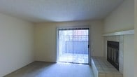 This single-story space presents a straightforward and functional layout with a clean, open living area. The room features wall-to-wall carpeting that adds a soft, comfortable feel underfoot, complemented by smooth, light-colored walls that create a neutral and adaptable interior palette. A textured popcorn ceiling runs throughout, a common characteristic of its era of construction.

A prominent sliding glass door serves as the focal point of the main living area, fitted with vertical blinds that allow for adjustable light control. The door provides direct access to an outdoor patio or enclosed balcony area, visible through the glass, offering a degree of natural light to the interior. The view through the door suggests a partially covered or fenced outdoor space.

Along one wall, a tiled step or raised platform detail is visible, finished in neutral square tiles, which may serve as a decorative ledge or functional surface. This architectural element adds a modest point of interest to an otherwise simply appointed room. Overall, the space presents a practical and move-in-ready environment well-suited for everyday living.
