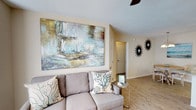 Located at 955 Airport Rd in the heart of Destin, Florida, this single-floor property offers a warm and inviting coastal retreat. The living area sets the tone for the entire space, showcasing light wood-style flooring that runs seamlessly underfoot, contributing to a bright and airy atmosphere. A large, eye-catching abstract canvas dominates one wall, featuring soft oceanic hues of blue, green, and gold that complement the coastal theme throughout. A comfortable neutral-toned sofa is adorned with coral and sea-themed accent pillows, reinforcing the beachy, relaxed ambiance. Decorative starburst wall accents add a stylish, mid-century coastal touch to the room. A ceiling fan overhead ensures comfortable airflow, ideal for the warm Florida climate. The space flows naturally toward additional rooms through a clean hallway, hinting at a well-organized and functional floor plan. The overall design of the property strikes a balance between casual beach living and tasteful interior styling, making it a comfortable and visually appealing space. Its single-floor layout ensures easy accessibility and a cohesive living experience throughout, well-suited for those seeking the relaxed charm of Destin's coastal lifestyle.