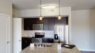 Located at The Vaughan in Austin, TX, this two-bedroom, two-bathroom apartment delivers a thoughtfully designed single-floor layout that balances style and functionality. The open-concept kitchen is a standout feature, showcasing rich dark cabinetry, granite countertops, a white subway tile backsplash, and a full suite of stainless steel appliances including a French door refrigerator, electric range, and built-in microwave. A large kitchen island with a built-in sink and pendant lighting serves as both a prep area and a natural gathering point, flowing seamlessly into the living area. The living room features plush carpeting, a ceiling fan, and multiple windows that invite natural light and provide outdoor views. Rich wood-look flooring runs through the kitchen and bathrooms, adding warmth and contrast. Both bedrooms are carpeted, well-sized, and include ceiling fans and windows with exterior views. The primary bathroom offers a granite-topped double vanity, wood-look flooring, a tub-shower combo with tile surround, and access to a walk-in closet. The second bathroom mirrors this clean aesthetic with a tub-shower combo and tile surround. Neutral wall tones throughout create a cohesive, move-in-ready ambiance.