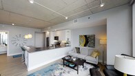 This 2-bedroom, 2-bathroom residence offers a thoughtfully designed open-concept floor plan that maximizes both space and functionality. The living and kitchen areas flow seamlessly together, anchored by striking exposed concrete ceilings that lend an industrial-modern character to the space. Track lighting runs along the ceiling, providing adjustable illumination throughout the main living area.

The kitchen is outfitted with crisp white cabinetry, stainless steel appliances including a refrigerator and microwave, and a dark stone countertop that extends into a breakfast bar — ideal for casual dining or entertaining. A tasteful tile backsplash adds texture and visual interest behind the range.

The adjacent living area is open and inviting, with warm hardwood-style flooring and ample room for comfortable furnishings. The space is brightened by recessed and ambient lighting, creating a welcoming atmosphere throughout the day and evening.

With two full bedrooms and two bathrooms, this residence provides a well-balanced layout suited for both individuals and roommates alike. The combination of modern fixtures, industrial design elements, and an efficient open floor plan makes this a highly functional and stylish urban living space.