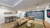 Located at 901 Leland St in Philadelphia's vibrant 19130 neighborhood, this well-appointed communal space offers a thoughtfully designed open-plan layout that seamlessly blends lounge, dining, and kitchenette areas within a single, cohesive floor. Light hardwood flooring runs throughout the interior, complemented by recessed lighting and crisp white ceilings that enhance the sense of brightness and openness.

The lounge area features a comfortable sofa, a rounded ottoman coffee table, and wire-frame accent tables arranged atop a green area rug. A wall-mounted flat-screen TV and a mid-century-style media console anchor one end of the room. Adjacent to the lounge, a compact kitchenette provides white cabinetry, a stainless steel refrigerator, a sink, and counter space, supported by open wooden shelving above. A high-top bar table and stools offer additional seating nearby.

A striking deep-blue accent wall defines the dining zone, enlivened by colorful framed artwork and a solid wood dining table with seating for several guests. Large floor-to-ceiling sliding glass doors and a secondary glass-paneled door connect the interior directly to a private outdoor patio. The patio features tile flooring, bold black-and-white striped ottomans, coral-red garden stools, a wood slat coffee table, and a shaded umbrella — creating an inviting extension of the interior living space.