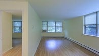 This single-floor residential space offers a straightforward and functional layout with a clean, understated aesthetic. The main living area is an open room finished with light hardwood flooring that reflects natural light beautifully, contributing to a bright and airy atmosphere. Walls are painted in a soft, neutral tone that enhances the sense of openness throughout the space.

A prominent set of large windows serves as a central feature of the living area, allowing generous amounts of natural light to fill the room while offering views of lush greenery outside. The windows are fitted with horizontal blinds for adjustable privacy and light control. A window-mounted air conditioning unit provides climate comfort during warmer months, while baseboard heating runs along the lower perimeter of the walls for warmth during cooler seasons.

The room's layout is versatile, providing ample open floor space that can accommodate a variety of furniture arrangements. Clean white baseboards and simple wall finishes give the interior a tidy, well-maintained appearance. The overall ambiance is calm and neutral, offering a blank canvas well-suited for personalizing to a range of interior styles. The space is practical, comfortable, and filled with the natural warmth of daylight.