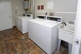 This single-floor laundry facility is designed with practicality and efficiency in mind, offering a communal space outfitted with commercial-grade Speed Queen washers and dryers. The room features exposed metallic ventilation ducts running from the machines up through the ceiling, reflecting the functional, utility-focused design of the space. An electrical panel is mounted on one wall alongside visible plumbing and exhaust connections, underscoring the room's purpose-built infrastructure. The walls are finished in a neutral tone and accented with posted signage — including notices reminding users to remove clothing promptly and informing them of management policies regarding lost or damaged articles. The layout is straightforward and accessible, with machines arranged for ease of use. The overall ambiance is clean and utilitarian, prioritizing convenience and shared use. The space is well-suited for residential or multi-unit building occupants seeking dependable on-site laundry access.