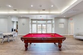 Located in Sun City West, Arizona, this well-appointed single-story community clubhouse offers a thoughtfully designed layout spanning a generous footprint. The main gathering area serves as the heart of the space, featuring a striking red-felt billiard table set beneath a tray ceiling with elegant cove lighting and recessed fixtures. Wood-look tile flooring flows throughout, lending a warm, cohesive aesthetic. This open area transitions seamlessly into a comfortable lounge zone furnished with a large sectional sofa, a wall-mounted TV, and a classic fireplace — creating an inviting setting for relaxation. A dedicated coffee and snack bar with granite countertops and wood cabinetry lines one wall, while dining tables and chairs offer additional seating for socializing. The exercise room is well-equipped with cardio machines including treadmills and an elliptical, free weights, and a wall mirror, all set against soft sage-green accent walls and light hardwood-style flooring. Natural light enters through shuttered windows. The office features built-in shelving stocked with books, a workstation, and a multifunction printer, making it a functional resource hub. A laundry room and connecting hallway complete the layout, ensuring full convenience for residents of this active community.