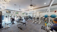 This fitness center in Houston, TX offers a comprehensive and well-appointed workout environment across a single, open floor plan. The space is designed with a clean, modern aesthetic, featuring crisp white walls accented by warm wood crown molding, patterned carpet tile flooring in cool blue-gray tones, and a combination of ceiling fans and distinctive spherical pendant light fixtures that provide ample illumination throughout.

The facility is organized into clearly defined workout zones. A dedicated cardio area houses a row of treadmills, elliptical machines, and stair climbers, each positioned for comfortable spacing. Multiple flat-screen televisions are mounted along the walls, allowing users to stay entertained during cardio sessions. Natural light filters in through glass-paneled doors and windows, adding brightness to the space.

The strength training area is equally well-equipped, featuring power racks, adjustable benches, a comprehensive dumbbell rack with a wide range of weights, cable machines, and various resistance equipment. Floor-to-ceiling mirrors line the walls, enhancing the sense of openness while allowing users to monitor their form.

A secondary room provides additional free weight and functional training space, complete with a water fountain, storage shelving, and a medicine ball rack. The overall ambiance is polished and motivating, reflecting a well-maintained, full-service fitness facility.