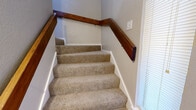 This efficiently designed 1-bedroom, 1-bathroom residence makes excellent use of its compact footprint, offering a well-organized layout that includes a combined living, dining, and kitchen area, a dedicated bedroom with closet, a full bathroom, and an interior staircase. The living room is a standout, featuring a classic brick fireplace, a ceiling fan, and a pass-through window opening to the kitchen, creating a warm and connected atmosphere. Soft carpet flooring runs throughout the main living spaces, complementing the neutral wall tones. A large sliding door with vertical blinds allows natural light to fill the room. The kitchen is fitted with rich wood cabinetry, light-toned countertops, a stainless steel sink, a dishwasher, and a black electric range with overhead ventilation. The bedroom is a comfortable size, also featuring a ceiling fan and access to a dedicated closet. The bathroom includes a tiled tub-and-shower combination, a wood-vanity with countertop, and light hardwood-style flooring. The carpeted staircase with a warm wood handrail adds a classic touch to the interior. Overall, the space presents a functional and inviting design with consistent finishes throughout.