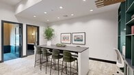 This thoughtfully designed single-floor amenity space offers a versatile blend of social and focused work environments. The main lounge area is anchored by a sleek, dark-topped communal bar table surrounded by plush olive-green upholstered barstools, set atop a soft, patterned area rug. Recessed lighting illuminates the crisp white walls, which are adorned with framed retro-inspired artwork adding a playful, creative touch to the atmosphere.

A standout teal built-in shelving unit with decorative accents and a slatted wood-and-metal ceiling detail creates a bold focal point, lending the space an artful, curated character. Adjacent to the main lounge, a cozy banquette dining area features a long tufted navy sofa flanked by warm wood-toned tables and leather accent chairs, all beneath a striking sculptural pendant light and a trio of large colorful framed prints.

Two private study rooms provide intimate, focused workspaces, each finished in warm wood paneling, deep navy carpet, a built-in white desk, wall sconce lighting, and a frosted window that filters natural light. Together, these zones create a well-rounded environment that balances collaborative gathering with individual productivity, all unified by a modern, design-forward aesthetic.