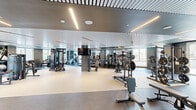 The fitness center at 4720 Third Ave is a generously sized, single-floor facility thoughtfully designed to accommodate a comprehensive range of workout needs. The space is divided into distinct functional zones, including a dedicated cardio area and a well-equipped strength training section, allowing users to move efficiently between activities.

The interior features a sleek, contemporary aesthetic defined by a distinctive ribbed ceiling with integrated linear LED lighting that evenly illuminates the floor below. The flooring transitions between rubberized surfaces in the free weights and strength areas and smooth dark flooring in the cardio zone, both chosen for durability and performance.

Large windows line the perimeter, flooding the space with natural light and offering open views of the surrounding neighborhood. Full-length mirrors along the walls enhance the sense of spaciousness while supporting proper form during training.

Equipment includes a row of treadmills, ellipticals, and stair climbers in the cardio zone, alongside cable machines, power racks, a dumbbell rack, and benches in the strength area. A wall-mounted television, water fountain, and restroom access add functional convenience. A bold graphic accent wall near the entrance provides a visual focal point, contributing to the facility's modern, energetic atmosphere.