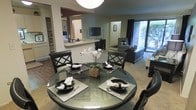 This single-floor residence showcases a thoughtfully designed open-concept layout that seamlessly connects the key living areas. The dining area serves as a central focal point, featuring a round glass-top table with elegant dark-framed chairs, set with coordinated place settings and accented by a fresh floral centerpiece. The space flows naturally into a pass-through kitchen, distinguished by a breakfast bar that provides both functional counter space and a visual transition between areas. The kitchen features light cabinetry and offers a peek into an adjacent room beyond. The living area extends comfortably from the dining space, furnished with a plush sofa and armchair arranged around an entertainment center housing a flat-screen television. A large sliding glass door dominates the far wall, flooding the living space with abundant natural light and offering a view of lush outdoor greenery, creating a seamless connection between the interior and exterior. Neutral wall tones and consistent flooring throughout the main areas lend the space a cohesive and inviting atmosphere. The combination of warm wood-toned flooring in the dining zone and carpeted living area adds textural variety, while the overall design reflects a clean, contemporary aesthetic ideal for both everyday living and entertaining.