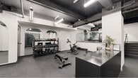 Located in the heart of Philadelphia's Rittenhouse Square neighborhood, this fitness facility offers a well-organized and comprehensive workout environment across a single open floor. The space features an industrial-chic aesthetic defined by exposed black ceiling beams, visible ductwork, and overhead linear lighting that creates a bold contrast against the clean white walls.

A standout architectural detail is the series of elegant arched mirror installations along one wall, which add visual depth and a refined character to the otherwise utilitarian space. The main workout area is equipped with a full rack of dumbbells and kettlebells, an adjustable weight bench, a stationary cycling bike, and a rowing machine. A dedicated cardio zone houses multiple treadmills positioned along a glass-railed partition.

A separate alcove provides additional functional training space, with wall-mounted equipment including gymnastic rings, foam rollers, and stability balls, creating a versatile area for bodyweight and mobility work. A reception or check-in counter with a sleek dark stone surface anchors the entry area, complemented by a storage unit for mats and accessories. The dark rubber flooring throughout is both durable and practical, completing the cohesive monochromatic design scheme of this efficient and modern fitness space.