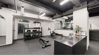 Located in the heart of Philadelphia's Rittenhouse Square neighborhood, this fitness facility offers a well-organized and comprehensive workout environment across a single open floor. The space features an industrial-chic aesthetic defined by exposed black ceiling beams, visible ductwork, and overhead linear lighting that creates a bold contrast against the clean white walls.

A standout architectural detail is the series of elegant arched mirror installations along one wall, which add visual depth and a refined character to the otherwise utilitarian space. The main workout area is equipped with a full rack of dumbbells and kettlebells, an adjustable weight bench, a stationary cycling bike, and a rowing machine. A dedicated cardio zone houses multiple treadmills positioned along a glass-railed partition.

A separate alcove provides additional functional training space, with wall-mounted equipment including gymnastic rings, foam rollers, and stability balls, creating a versatile area for bodyweight and mobility work. A reception or check-in counter with a sleek dark stone surface anchors the entry area, complemented by a storage unit for mats and accessories. The dark rubber flooring throughout is both durable and practical, completing the cohesive monochromatic design scheme of this efficient and modern fitness space.