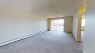 This single-floor, one-bedroom, one-bathroom unit offers a straightforward and functional layout designed for comfortable everyday living. The living area is generously sized, featuring neutral gray carpeting that extends throughout and contributes to a clean, understated aesthetic. White walls provide a blank canvas, making the space easy to personalize. A large sliding glass door dominates one wall of the living area, fitted with vertical blinds that allow for adjustable light control. The door provides an abundance of natural light, brightening the interior throughout the day. A baseboard heating unit runs along the perimeter wall, offering an efficient and consistent heat source during colder months. The textured ceiling adds a subtle architectural detail consistent with the unit's practical design. The overall ambiance is calm and neutral, with a simple, livable character that suits a range of furnishing styles. The combination of ample natural light, open floor space, and functional heating infrastructure makes this a practical and versatile living space well-suited for a single occupant or couple.