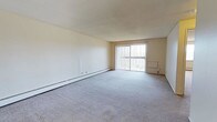 This single-floor, one-bedroom, one-bathroom unit offers a straightforward and functional layout designed for comfortable everyday living. The living area is generously sized, featuring neutral gray carpeting that extends throughout and contributes to a clean, understated aesthetic. White walls provide a blank canvas, making the space easy to personalize. A large sliding glass door dominates one wall of the living area, fitted with vertical blinds that allow for adjustable light control. The door provides an abundance of natural light, brightening the interior throughout the day. A baseboard heating unit runs along the perimeter wall, offering an efficient and consistent heat source during colder months. The textured ceiling adds a subtle architectural detail consistent with the unit's practical design. The overall ambiance is calm and neutral, with a simple, livable character that suits a range of furnishing styles. The combination of ample natural light, open floor space, and functional heating infrastructure makes this a practical and versatile living space well-suited for a single occupant or couple.