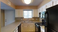 This single-story space includes a kitchen designed for efficient use of space. The kitchen features white cabinetry that lines the walls, offering ample storage through both upper and lower cabinets. The countertops provide a neutral, light-toned workspace that complements the overall color palette. Black appliances, including an electric range and an over-the-range microwave, add a practical contrast to the white cabinetry. A double-basin sink is positioned beneath a window, allowing natural light to fill the space while offering an outdoor view during meal preparation. The warm beige wall tones contribute to a welcoming atmosphere throughout. A flush-mount ceiling light fixture ensures consistent, even illumination across the kitchen. The layout follows a U-shaped configuration, maximizing workflow efficiency and providing easy access to all areas of the kitchen. A dishwasher is also integrated into the design, further adding to the functionality of the space. The overall aesthetic is clean and straightforward, making it a practical and comfortable kitchen environment.