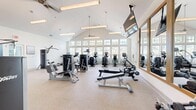 Located at 2525 Sycamore Farms Dr NW in Concord, NC, this fitness center offers a thoughtfully designed workout environment on a single floor. The space is defined by its striking vaulted ceiling, which soars overhead and contributes to an open, airy atmosphere. A generous row of large, multi-pane windows lines the perimeter, flooding the interior with natural light and providing views of the surrounding outdoor area, including what appears to be a pool and landscaped grounds.

The fitness center is outfitted with a comprehensive suite of Life Fitness equipment, including stair climbers, elliptical machines, treadmills, and a variety of strength-training and cable machines. A dedicated free weights area features a dumbbell rack accompanied by wall-mounted mirrors and flat-screen TVs, adding convenience and motivation during workouts. Ceiling fans and pendant lighting fixtures complement the vaulted roofline, enhancing both comfort and aesthetics.

The flooring throughout is a durable, speckled rubberized surface, well-suited for a high-use fitness environment. Floor-to-ceiling glass panel walls and doors on one side of the space provide a seamless visual connection to an adjacent area, adding to the sense of openness. The overall design is clean, modern, and bright, creating an inviting and functional space for a full range of fitness activities.