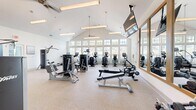 Located at 2525 Sycamore Farms Dr NW in Concord, NC, this fitness center offers a thoughtfully designed workout environment on a single floor. The space is defined by its striking vaulted ceiling, which soars overhead and contributes to an open, airy atmosphere. A generous row of large, multi-pane windows lines the perimeter, flooding the interior with natural light and providing views of the surrounding outdoor area, including what appears to be a pool and landscaped grounds.

The fitness center is outfitted with a comprehensive suite of Life Fitness equipment, including stair climbers, elliptical machines, treadmills, and a variety of strength-training and cable machines. A dedicated free weights area features a dumbbell rack accompanied by wall-mounted mirrors and flat-screen TVs, adding convenience and motivation during workouts. Ceiling fans and pendant lighting fixtures complement the vaulted roofline, enhancing both comfort and aesthetics.

The flooring throughout is a durable, speckled rubberized surface, well-suited for a high-use fitness environment. Floor-to-ceiling glass panel walls and doors on one side of the space provide a seamless visual connection to an adjacent area, adding to the sense of openness. The overall design is clean, modern, and bright, creating an inviting and functional space for a full range of fitness activities.