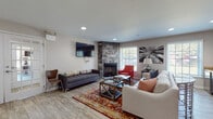 This well-appointed single-floor space combines modern design with warm, inviting elements to create a comfortable and functional environment. The centerpiece of the main living area is a striking floor-to-ceiling stone corner fireplace, which anchors the room and adds a rustic yet refined character. Light wood-look flooring flows throughout, complementing the neutral wall tones and creating a bright, cohesive aesthetic.

The living area is generously sized, accommodating multiple seating arrangements including a plush sectional sofa, a tufted daybed, and a bold red accent chair that adds a pop of color. Recessed lighting overhead, paired with large windows dressed in white horizontal blinds, ensures the space is well-lit with ample natural light and outdoor views. A wall-mounted flat-screen television provides a central entertainment focal point.

Seamlessly connected to the living area is a built-in kitchenette, featuring dark espresso cabinetry, granite countertops, and a utility sink — ideal for refreshments and light food preparation. A vibrant burnt-orange accent wall nearby adds a dynamic design element, with French doors leading to an adjacent area.

A separate dining nook offers a tall dark wood table with leather stools and additional seating, making the space well-suited for gatherings and collaborative use.