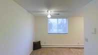 This single-floor space offers a clean, functional layout with a welcoming ambiance. The main room is characterized by neutral cream-toned walls that create a bright, open atmosphere, complemented by wall-to-wall carpeting that adds warmth underfoot. A ceiling fan with an integrated light fixture is centrally positioned, providing both air circulation and overhead lighting throughout the room. A window fitted with horizontal blinds allows natural light to filter in while offering a view of the surrounding tree line, connecting the interior to the outdoors. A baseboard heating unit runs along the exterior wall beneath the window, ensuring efficient climate control throughout the colder months. The textured ceiling adds a classic finish to the space. Overall, the room presents a straightforward, versatile layout well-suited for a variety of uses, with practical features that prioritize comfort and functionality. The simplicity of the design allows for flexible furnishing and personalization to suit a range of needs.