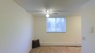 This single-floor space offers a clean, functional layout with a welcoming ambiance. The main room is characterized by neutral cream-toned walls that create a bright, open atmosphere, complemented by wall-to-wall carpeting that adds warmth underfoot. A ceiling fan with an integrated light fixture is centrally positioned, providing both air circulation and overhead lighting throughout the room. A window fitted with horizontal blinds allows natural light to filter in while offering a view of the surrounding tree line, connecting the interior to the outdoors. A baseboard heating unit runs along the exterior wall beneath the window, ensuring efficient climate control throughout the colder months. The textured ceiling adds a classic finish to the space. Overall, the room presents a straightforward, versatile layout well-suited for a variety of uses, with practical features that prioritize comfort and functionality. The simplicity of the design allows for flexible furnishing and personalization to suit a range of needs.