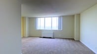 This single-floor space presents a clean, functional layout centered around a generously sized main living area. The room is finished with soft wall-to-wall carpeting underfoot, lending a warm and quiet atmosphere throughout. Pale yellow walls contribute a light, neutral tone that complements the natural brightness of the space. The most striking architectural feature is the wide, multi-pane window spanning a substantial portion of one wall, framing broad outdoor views and drawing in abundant natural light. Sheer curtains flank the window, offering privacy while preserving the airy feel of the room. A white baseboard heating unit is centrally positioned beneath the window, integrating practical climate control seamlessly into the design. The ceiling is smooth and unadorned, reinforcing the room's understated, minimalist character. Corners and transitions between walls are cleanly finished, reflecting a straightforward and well-maintained interior. The open floor plan allows for flexible furniture arrangement, making the space highly adaptable to a variety of uses. Overall, the space offers a bright, calm environment with a focus on comfort and practicality.