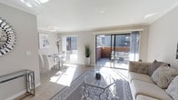 This stylish single-floor space is defined by its clean, contemporary design and an abundance of natural light. The open-plan layout seamlessly connects the living and dining areas, creating a cohesive and inviting atmosphere throughout. Neutral tones — soft whites, warm grays, and light wood-effect flooring — establish a calm, sophisticated palette that runs consistently across the space.

The dining area features a sleek glass-top table paired with modern white chairs, positioned near a large window that draws in plentiful daylight. Tasteful wall art and a striking floral arrangement add curated elegance to the corner. The living area is anchored by a glass coffee table set atop a patterned area rug, complemented by plush neutral seating that reinforces the space's refined aesthetic.

A standout feature is the expansive sliding glass door that opens directly onto a private outdoor terrace. The terrace, framed by brick boundary walls, is furnished with comfortable outdoor seating, offering a seamless indoor-outdoor living experience with open views of the surrounding neighborhood. Sheer drapes frame the glass doors, softening the transition between interior and exterior. Altogether, the space strikes a balance between modern minimalism and warm livability.