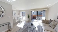 This stylish single-floor space is defined by its clean, contemporary design and an abundance of natural light. The open-plan layout seamlessly connects the living and dining areas, creating a cohesive and inviting atmosphere throughout. Neutral tones — soft whites, warm grays, and light wood-effect flooring — establish a calm, sophisticated palette that runs consistently across the space.

The dining area features a sleek glass-top table paired with modern white chairs, positioned near a large window that draws in plentiful daylight. Tasteful wall art and a striking floral arrangement add curated elegance to the corner. The living area is anchored by a glass coffee table set atop a patterned area rug, complemented by plush neutral seating that reinforces the space's refined aesthetic.

A standout feature is the expansive sliding glass door that opens directly onto a private outdoor terrace. The terrace, framed by brick boundary walls, is furnished with comfortable outdoor seating, offering a seamless indoor-outdoor living experience with open views of the surrounding neighborhood. Sheer drapes frame the glass doors, softening the transition between interior and exterior. Altogether, the space strikes a balance between modern minimalism and warm livability.