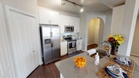 This single-story, two-bedroom, two-bathroom residence offers a well-appointed layout across a generously sized floor plan. The kitchen serves as a functional centerpiece, featuring stainless steel appliances including a side-by-side refrigerator with ice and water dispenser, an electric range, and a built-in microwave. White shaker-style cabinetry extends to the ceiling, complemented by gray quartz countertops, a subway tile backsplash, and dark hardwood-style flooring. Track lighting illuminates the space, and an elegant arched passageway connects the kitchen to the adjacent living areas. A curved breakfast bar provides additional counter seating and opens toward the carpeted living room, which benefits from natural light and ample storage via double-door closets. Both bedrooms are carpeted and include ceiling fans with lighting, as well as windows with blinds that allow natural light. The primary bedroom features vaulted ceilings and multiple windows. Both bathrooms are finished with gray quartz countertops, white cabinetry, undermount sinks, and tub-shower combinations with neutral tile surround. The overall aesthetic is clean, modern, and cohesive throughout.