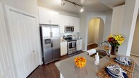 This single-story, two-bedroom, two-bathroom residence offers a well-appointed layout across a generously sized floor plan. The kitchen serves as a functional centerpiece, featuring stainless steel appliances including a side-by-side refrigerator with ice and water dispenser, an electric range, and a built-in microwave. White shaker-style cabinetry extends to the ceiling, complemented by gray quartz countertops, a subway tile backsplash, and dark hardwood-style flooring. Track lighting illuminates the space, and an elegant arched passageway connects the kitchen to the adjacent living areas. A curved breakfast bar provides additional counter seating and opens toward the carpeted living room, which benefits from natural light and ample storage via double-door closets. Both bedrooms are carpeted and include ceiling fans with lighting, as well as windows with blinds that allow natural light. The primary bedroom features vaulted ceilings and multiple windows. Both bathrooms are finished with gray quartz countertops, white cabinetry, undermount sinks, and tub-shower combinations with neutral tile surround. The overall aesthetic is clean, modern, and cohesive throughout.