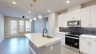 This well-designed 2-bedroom, 2-bathroom residence offers a thoughtful single-floor layout that maximizes comfort and functionality. The open-concept kitchen and living area serves as the heart of the home, anchored by a large center island with a white quartz countertop and a matte black gooseneck faucet. The kitchen is outfitted with crisp white cabinetry, a dark linear tile backsplash, and a full suite of stainless steel appliances, including a gas range, built-in microwave, and French door refrigerator. Globe pendant lights with brass accents add a contemporary touch overhead. Light wood-look flooring flows throughout the space, complementing the neutral warm-toned walls. The living area features a ceiling fan, abundant natural light from multiple windows, and direct outdoor access through a glass-panel door. The primary bedroom is generously proportioned and includes its own en-suite bathroom, which boasts a dual-sink vanity with white cabinetry, matte black fixtures, a large framed mirror, and a glass-enclosed shower. A spacious walk-in closet with wrap-around shelving provides ample storage. The second bedroom is equally bright, with windows and a ceiling fan. A hallway connects the private spaces efficiently, completing this clean, modern layout.