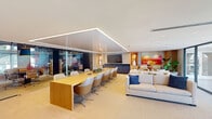 Located at 1000 Seminole Drive in Fort Lauderdale, FL, this single-floor professional office suite offers a thoughtfully designed, open-concept workspace that seamlessly blends collaborative and private environments. The main area is anchored by a substantial natural wood conference table surrounded by modern ergonomic seating, positioned beneath a striking geometric recessed ceiling detail with integrated linear LED lighting that adds both warmth and architectural interest. Adjacent to the central workspace, a comfortable lounge area features plush neutral sofas accented with bold blue throw pillows, set against a backdrop of vibrant, large-scale abstract artwork. A sleek media wall with built-in display and warm accent niches adds refinement to the space. A series of glass-walled private conference rooms lined along one side offer full visibility while maintaining acoustic separation, sliding on modern hardware tracks. One intimate conference room stands out with a dramatic full-wall mural, deep blue ceiling, statement pendant lighting, and a round dark table with classic leather task chairs. Floor-to-ceiling glazing along the perimeter floods the space with natural light and provides open views of the surrounding Fort Lauderdale landscape, further enhancing the bright, airy atmosphere throughout. A kitchenette area completes the suite's functional offerings.