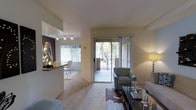 This single-floor residence offers a thoughtfully designed open-concept layout that seamlessly connects the living, dining, and kitchen areas. The living space is anchored by plush wall-to-wall carpeting and is bathed in natural light, with large sliding glass doors that open directly onto a private balcony overlooking lush greenery. The balcony provides a tranquil outdoor retreat with pleasant tree-lined views, blurring the boundary between indoor and outdoor living. Adjacent to the living area, a raised breakfast bar with a dark granite countertop defines the kitchen boundary and adds a stylish, functional element to the space. The dining nook, situated near a window with bright, filtered light, provides an intimate setting for meals. Track lighting on the ceiling accents the kitchen and dining areas with warm, focused illumination. The neutral, warm-toned walls throughout lend the space a calm and versatile ambiance, while decorative touches — including sculptural art pieces and layered rugs — add personality and depth. Overall, the layout is practical yet inviting, offering a comfortable and well-lit environment with appealing outdoor connectivity.