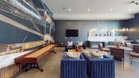 Located at 143 E White St in Rock Hill, SC, this single-floor amenity lounge is designed with a modern industrial aesthetic that blends bold style with functional recreation. The space is anchored by deep navy-blue walls, polished concrete floors, and a dramatic oversized photographic mural depicting industrial machinery, lending the interior a distinctive, urban character.

The main area features a regulation ping pong table set atop a geometric patterned area rug, positioned beneath a sculptural multi-arm pendant light fixture. Large floor-to-ceiling windows and an oversized garage-style door flood the space with natural light and provide views of an outdoor area, creating a seamless indoor-outdoor connection.

A dedicated lounge zone offers channeled navy velvet seating, cognac leather accent chairs, and a shuffleboard table, all set against the striking industrial mural wall. A wall-mounted television is positioned nearby, alongside a small beverage and snack bar area with open shelving. An additional seating cluster with a round table provides a more intimate setting. Curated artwork and layered lighting complete the sophisticated, community-focused ambiance of this well-appointed gathering space.