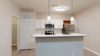 This single-floor residence offers a thoughtfully designed open-concept layout that seamlessly connects the kitchen and living areas. The kitchen is a standout feature, equipped with stainless steel appliances including a refrigerator, microwave, and range, all complemented by crisp white cabinetry, a white subway tile backsplash, and a light-toned countertop peninsula. Pendant lighting and a flush ceiling fixture illuminate the space with warm, focused light, while a built-in wine rack adds a charming detail. Light wood-style flooring runs throughout the main living and kitchen areas, contributing to a bright and cohesive aesthetic. The living area is generously proportioned, featuring a ceiling fan with light fixture and neutral walls that create a versatile, move-in-ready canvas. A window allows natural light to filter in, enhancing the open feel. The bedroom continues the warm, neutral palette with plush carpeting, a ceiling fan, and a window overlooking greenery, providing a comfortable and serene retreat with closet access. The bathroom is finished with a white vanity, light countertop, a tiled tub-and-shower combination, and wood-style flooring, offering a clean and functional design throughout.