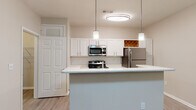 This single-floor residence offers a thoughtfully designed open-concept layout that seamlessly connects the kitchen and living areas. The kitchen is a standout feature, equipped with stainless steel appliances including a refrigerator, microwave, and range, all complemented by crisp white cabinetry, a white subway tile backsplash, and a light-toned countertop peninsula. Pendant lighting and a flush ceiling fixture illuminate the space with warm, focused light, while a built-in wine rack adds a charming detail. Light wood-style flooring runs throughout the main living and kitchen areas, contributing to a bright and cohesive aesthetic. The living area is generously proportioned, featuring a ceiling fan with light fixture and neutral walls that create a versatile, move-in-ready canvas. A window allows natural light to filter in, enhancing the open feel. The bedroom continues the warm, neutral palette with plush carpeting, a ceiling fan, and a window overlooking greenery, providing a comfortable and serene retreat with closet access. The bathroom is finished with a white vanity, light countertop, a tiled tub-and-shower combination, and wood-style flooring, offering a clean and functional design throughout.