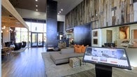 This striking single-story space makes an immediate impression with its soaring double-height ceilings and bold, contemporary design aesthetic. The focal point of the main area is a floor-to-ceiling reclaimed wood accent wall, adding warmth and texture that contrasts beautifully against the dark, industrial-style finishes throughout. A sleek, modern double-sided fireplace anchored by a stacked log display serves as a dramatic architectural centerpiece, blending function with visual impact.

The seating area features a low-profile sectional sofa in neutral tones, accented with vibrant orange throw pillows that inject energy into the otherwise cool palette. A chunky wooden cube side table and a plush area rug ground the lounge space, creating an inviting and relaxed atmosphere. Large glass entry doors flood the interior with natural light and offer views to an outdoor courtyard or patio area beyond.

Dark hardwood flooring runs throughout, complementing the space's industrial-modern character. Recessed lighting illuminates the interior thoughtfully, while framed artwork and a digital display screen add refined, curated details. The overall ambiance is sophisticated, open, and welcoming.