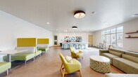 This single-floor community room in Madison, WI offers a generously sized, open-plan layout designed to accommodate a variety of social and recreational activities. The space is finished with large-format tile flooring throughout, crisp white walls, and recessed lighting complemented by modern pendant fixtures, creating a bright and welcoming atmosphere.

A standout feature is the long, white marble-topped bar counter, lined with green stools and backed by a bold geometric teal-and-white tile accent wall. A coffee and beverage station adds functional appeal to this central gathering point. Pendant lights hang above the counter, enhancing both form and function.

The lounge area is defined by a wall-mounted flat-screen TV above a natural wood console table, with a round upholstered ottoman providing casual seating. A colorful abstract painting and high-top bistro table with metal barstools add character to this corner. A vibrant yellow-green privacy booth near the entrance offers a semi-enclosed nook for quieter use.

Large windows and glass doors along one wall flood the space with natural light and provide views of the surrounding neighborhood, while double entry doors with glass panels ensure easy accessibility throughout.
