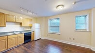 This efficiently designed one-bedroom, one-bathroom unit is laid out across a single floor, making smart use of every square foot. Rich, dark hardwood flooring runs throughout the main living areas, adding warmth and character to the space.

The kitchen is well-appointed with warm oak cabinetry extending both above and below the countertops, offering generous storage. It includes a white electric range, a refrigerator, a double-basin stainless steel sink, and dark granite-style countertops. Track lighting overhead ensures the kitchen is well-lit and functional.

The living and dining area flows naturally from the kitchen, benefiting from a window fitted with blinds that lets in natural light while maintaining privacy. A ceiling fan is present in the bedroom, which features two windows that further brighten the room.

The bathroom is clean and well-finished, with classic white hex tile flooring, a full tub-shower combination with white tile surround accented by dark border tiles, a light wood vanity with a marble-style countertop, and a mirrored medicine cabinet above. The overall palette of soft cream walls, white trim, and hardwood floors gives the unit a cohesive, inviting feel throughout.