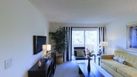 This single-floor space offers a comfortable and contemporary living environment designed with both style and functionality in mind. The living room serves as the centerpiece of the home, characterized by neutral wall tones and soft carpeted flooring that create a warm, inviting atmosphere. A large sliding glass door floods the room with natural light and provides direct views of lush greenery outside, giving the space an airy, open feel. Bold black-and-white striped curtains frame the door, adding a striking design contrast to the otherwise understated color palette. The living area is thoughtfully arranged with modern, dark-toned furniture, including a low-profile media console that anchors a wall-mounted flat-screen television. A sleek coffee table and coordinating seating complete the arrangement, while decorative accents — including potted plants, floral arrangements, and ambient table lamps — add personality and warmth to the room. The smooth, light-toned ceiling contributes to the sense of openness throughout the space. Overall, the home presents a clean, modern aesthetic with a well-balanced mix of functional design and decorative detail, making it a comfortable and visually appealing living environment.
