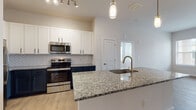 This single-floor residence offers a thoughtfully designed open layout with contemporary finishes throughout. The kitchen serves as a focal point, showcasing granite countertops atop a two-tone cabinet scheme of crisp white uppers and deep navy lowers. A large center island with an undermount sink and pendant lighting provides additional workspace and casual seating. Stainless steel appliances, including a range and over-the-range microwave, are set against a herringbone-patterned tile backsplash. Light wood-look flooring runs throughout the main living areas, connecting the kitchen to a spacious living area with large windows and a door that allows abundant natural light to fill the space.

The bedrooms feature plush carpet flooring, ceiling fans, and windows fitted with blinds. The primary bedroom connects directly to a private en-suite bathroom with a dual-sink granite vanity, walk-in closet, and a separate shower area. A secondary bathroom offers a tiled tub-and-shower combination with brushed nickel fixtures and a granite-topped vanity. The overall aesthetic is clean and modern, with neutral tones and quality finishes throughout.
