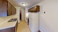 This single-floor, 1-bedroom, 1-bathroom unit offers a straightforward and functional layout designed for comfortable everyday living. The living room is generously proportioned, featuring wall-to-wall carpeting, neutral cream-toned walls, and a textured ceiling. A large sliding glass door floods the space with natural light and provides direct views of the surrounding greenery, while a baseboard heating unit and wall-mounted air conditioner ensure year-round climate control. The entry door, finished in warm dark wood, adds a classic touch to the space.

The galley-style kitchen is efficiently arranged, offering dark wood cabinetry for ample storage, a double-basin sink with light countertops, and white appliances including a refrigerator and electric range. Tile flooring and a flush-mount ceiling light complete the kitchen's practical design. A hallway from the kitchen connects to the rest of the unit.

The bedroom continues the carpeted flooring and neutral palette, with a wide sliding window that frames a tranquil view of the outdoor landscape, including open lawn and surrounding trees. A baseboard heater runs beneath the window.

The bathroom features classic white square wall tiling surrounding a full soaking tub with shower, a mosaic tile floor, a vanity with storage, and a dark wood door — offering a clean, timeless aesthetic throughout.