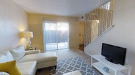 This two-story residence offers a welcoming main living area characterized by warm yellow-toned walls, plush carpeting, and a textured ceiling that adds subtle visual interest. The focal point of the living room is a full-width sliding glass door dressed with vertical blinds, allowing ample natural light to fill the space while providing direct access to an outdoor patio area, where greenery is visible beyond. A decorative white metal staircase railing featuring an elegant branch-inspired pattern serves as a standout architectural detail, adding an artistic touch to the transition between floors. The staircase leads to the second level, suggesting additional living or sleeping quarters above. The living room is generously proportioned, easily accommodating a seating arrangement anchored by a patterned area rug. A media console positioned near the staircase provides a practical entertainment setup. Subtle recessed ventilation and thoughtful layout details reflect a functional yet comfortable design. Overall, the space presents a bright, casual ambiance suited for everyday living, with natural light and outdoor connectivity as defining characteristics.