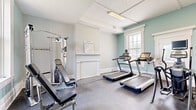 The District Fit Studio is a thoughtfully designed fitness facility housed within a classically styled building. The entrance welcomes visitors through a bright, airy foyer featuring light hardwood-style flooring, tall windows that invite natural light, and a branded welcome mat beneath an elegant gold ring chandelier. A staircase with warm wood treads, a dark metal banister, and a bold teal accent wall with a starburst detail adds a striking visual element to the hallway connecting the two exercise rooms.

The first exercise room is dedicated to cardiovascular and strength training, featuring multiple treadmills, a multi-function cable weight machine, and additional resistance equipment. Soft teal-painted walls complement light grey tones, while a large window provides natural light and ventilation. A traditional-style wainscoting detail adds architectural character.

The second exercise room focuses on cycling and cross-training, housing spin bikes, a rowing machine, a recumbent bike, and a StairMaster. Full-length wall mirrors expand the sense of space and allow exercisers to monitor their form. Bright overhead lighting and generous windows ensure a well-lit, energizing environment throughout.

Both rooms feature durable rubber-style flooring suited for fitness use, and the overall aesthetic balances classic architectural detailing with a clean, modern fitness atmosphere.