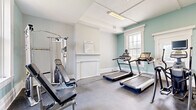 The District Fit Studio is a thoughtfully designed fitness facility housed within a classically styled building. The entrance welcomes visitors through a bright, airy foyer featuring light hardwood-style flooring, tall windows that invite natural light, and a branded welcome mat beneath an elegant gold ring chandelier. A staircase with warm wood treads, a dark metal banister, and a bold teal accent wall with a starburst detail adds a striking visual element to the hallway connecting the two exercise rooms.

The first exercise room is dedicated to cardiovascular and strength training, featuring multiple treadmills, a multi-function cable weight machine, and additional resistance equipment. Soft teal-painted walls complement light grey tones, while a large window provides natural light and ventilation. A traditional-style wainscoting detail adds architectural character.

The second exercise room focuses on cycling and cross-training, housing spin bikes, a rowing machine, a recumbent bike, and a StairMaster. Full-length wall mirrors expand the sense of space and allow exercisers to monitor their form. Bright overhead lighting and generous windows ensure a well-lit, energizing environment throughout.

Both rooms feature durable rubber-style flooring suited for fitness use, and the overall aesthetic balances classic architectural detailing with a clean, modern fitness atmosphere.