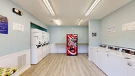 This laundry facility at 3940 Macon Road in Columbus, Georgia, offers a clean, efficiently organized space designed to accommodate all laundry needs with ease. The room is finished with light wood-look flooring and soft sage-green walls accented by white wainscoting, creating a bright and welcoming atmosphere. Overhead fluorescent lighting ensures the space is well-lit throughout.

Along one wall, a row of top-load washing machines provides ample capacity for multiple users, while a bank of stacked washer-dryer units lines the adjacent wall, maximizing the use of available space. Laminate folding counters are conveniently positioned near the machines, offering practical surfaces for sorting and folding laundry.

A padded bench seat is situated near the entrance, providing a comfortable waiting area for patrons. A Coca-Cola vending machine adds a convenient amenity for visitors during their stay. The entrance features a glass-panel door flanked by two windows, allowing natural light to filter in and offering views of the surrounding outdoor greenery.

Safety features include a mounted fire extinguisher and clearly posted usage instructions and informational signage. The space is well-maintained and thoughtfully arranged to provide a functional, user-friendly laundry experience.