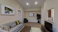 This single-floor residence offers a comfortable and thoughtfully designed living environment that balances style with functionality. The main living area serves as the centerpiece of the space, featuring warm neutral wall tones that create a calm and inviting atmosphere. Plush wall-to-wall carpeting adds softness underfoot, while recessed ceiling lighting provides even, ambient illumination throughout the room. The living area is generously proportioned, offering ample space for seating and entertaining. A gray sofa accented with green and patterned throw pillows pairs with a dark accent chair, anchoring the room around a rich wood coffee table and a patterned area rug. A flat-screen television is positioned on a sleek media console, making the space ideal for both relaxing and hosting. Framed wall art and a large potted indoor plant add natural and decorative touches that elevate the overall aesthetic. Multiple doorways lead to adjoining rooms, suggesting a well-connected and functional floor plan. The high ceilings enhance the sense of openness, contributing to an airy feel. The overall design reflects a clean, modern sensibility with warm, livable accents throughout.